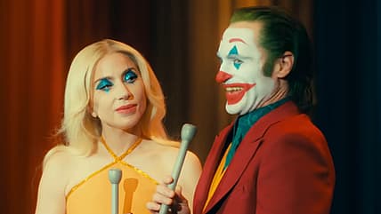 Arthur (Joaquin Phoenix) invites Harleen (Lady Gaga) to introduce their next bit in Joker: Folie à