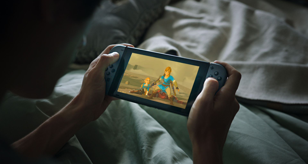 A man plays with a Nintendo Switch with grey Joy-Cons in bed. The Legend of Zelda: Breath of the Wild can be seen on the screen via Nintendo