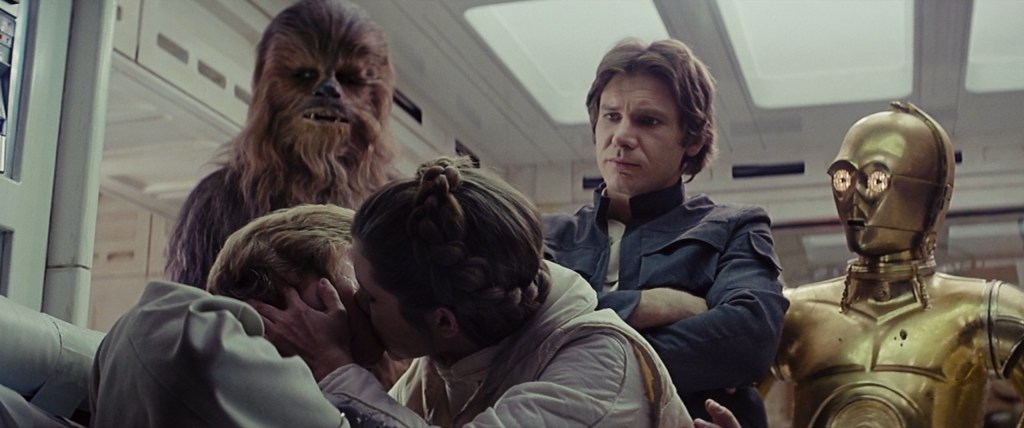 Princess Leia (Carrie Fisher) kisses Luke Skywalker (Mark Hamill) in front of Chewbacca (Peter Mayhew), Han Solo (Harrison Ford), and C-3PO (Anthony Daniels) in Star Wars Episode V: The Empire Strikes Back (1980), Lucasfilm Ltd.